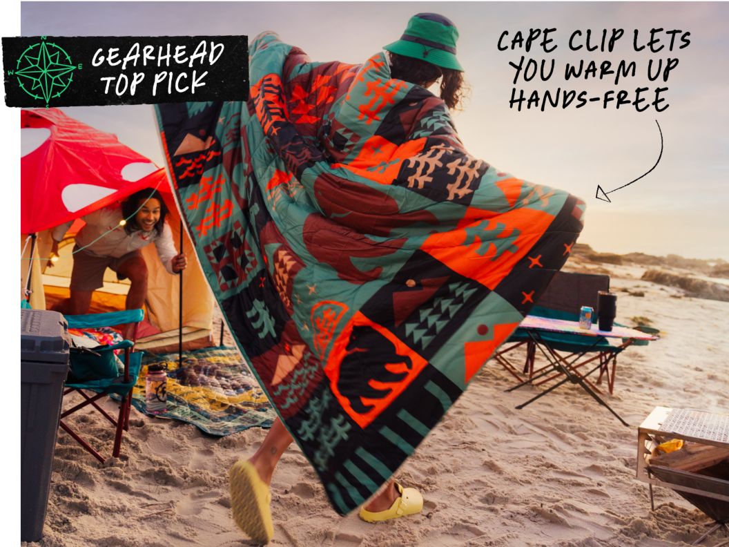 A person wearing a blanket at a beach campsite pretends that it is her wings. Text overlay reads: Gearhead top pick, cape cliip lets you warm up hands-free.